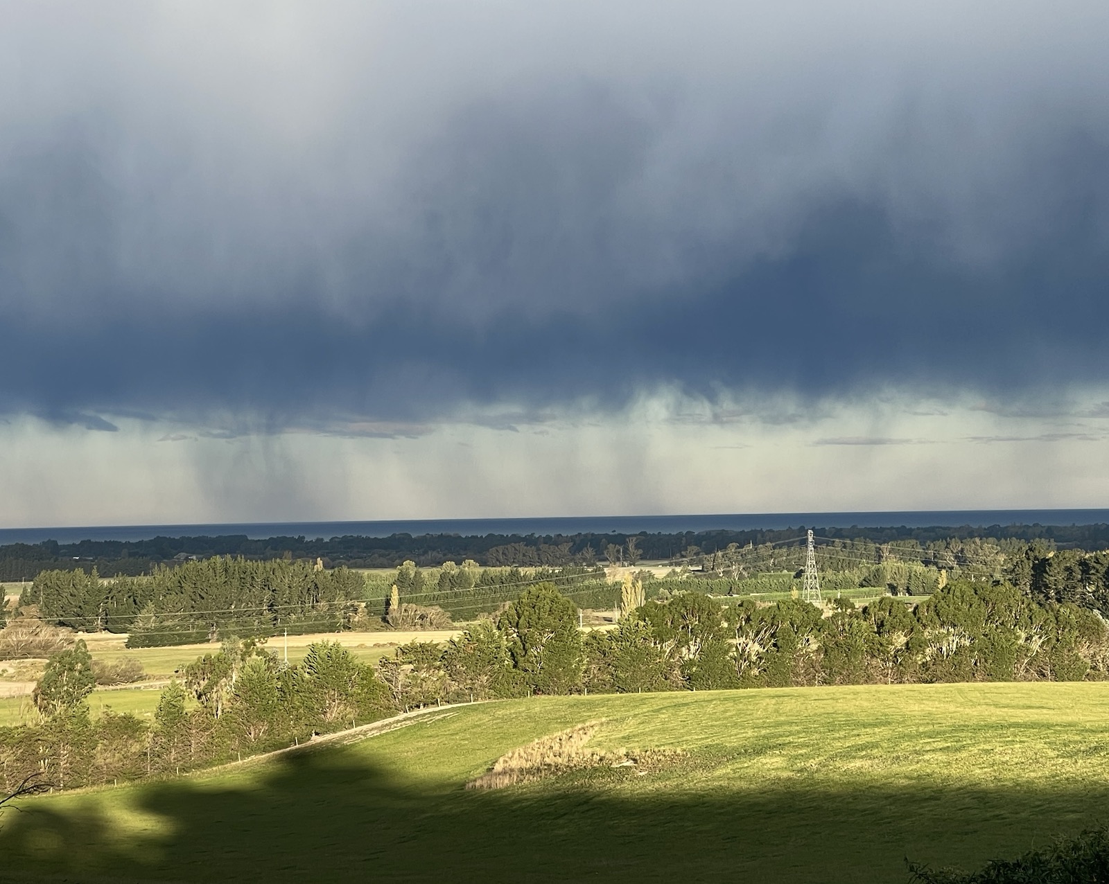 A dark rain squall spreading in a flat grey sheet across a wide green plain.