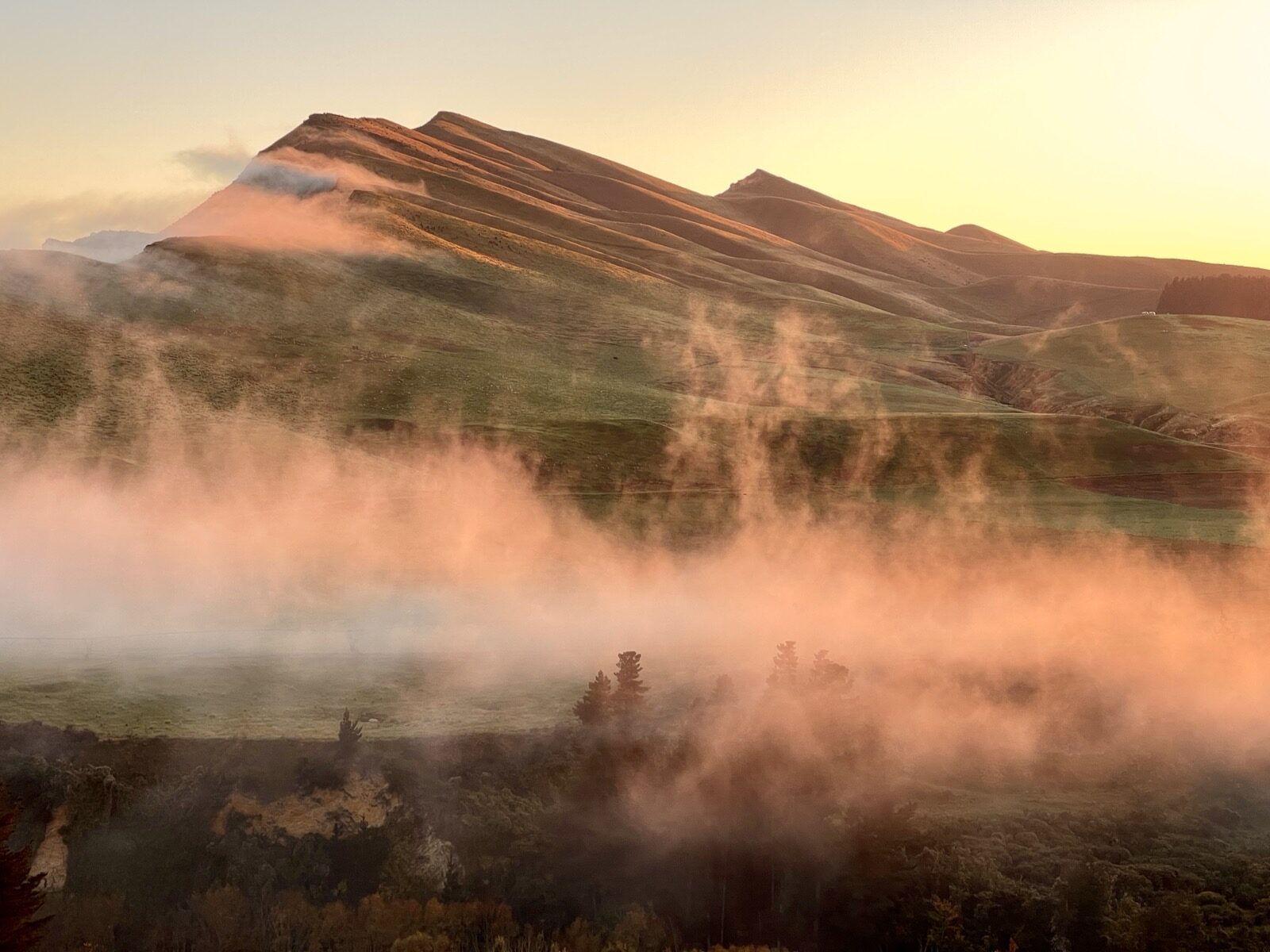 Morning mist drifting over a ridged hillside in warm low light, with wisps rising from the valley floor.