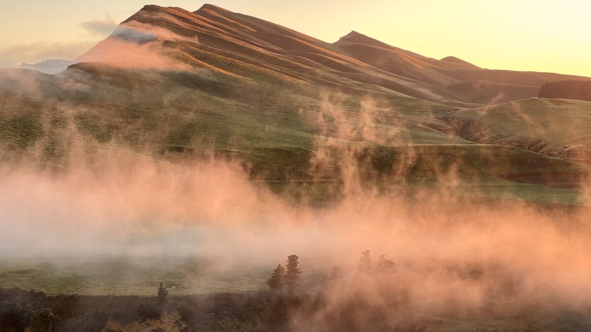 Morning mist drifting over a ridged hillside in warm low light, with wisps rising from the valley floor.